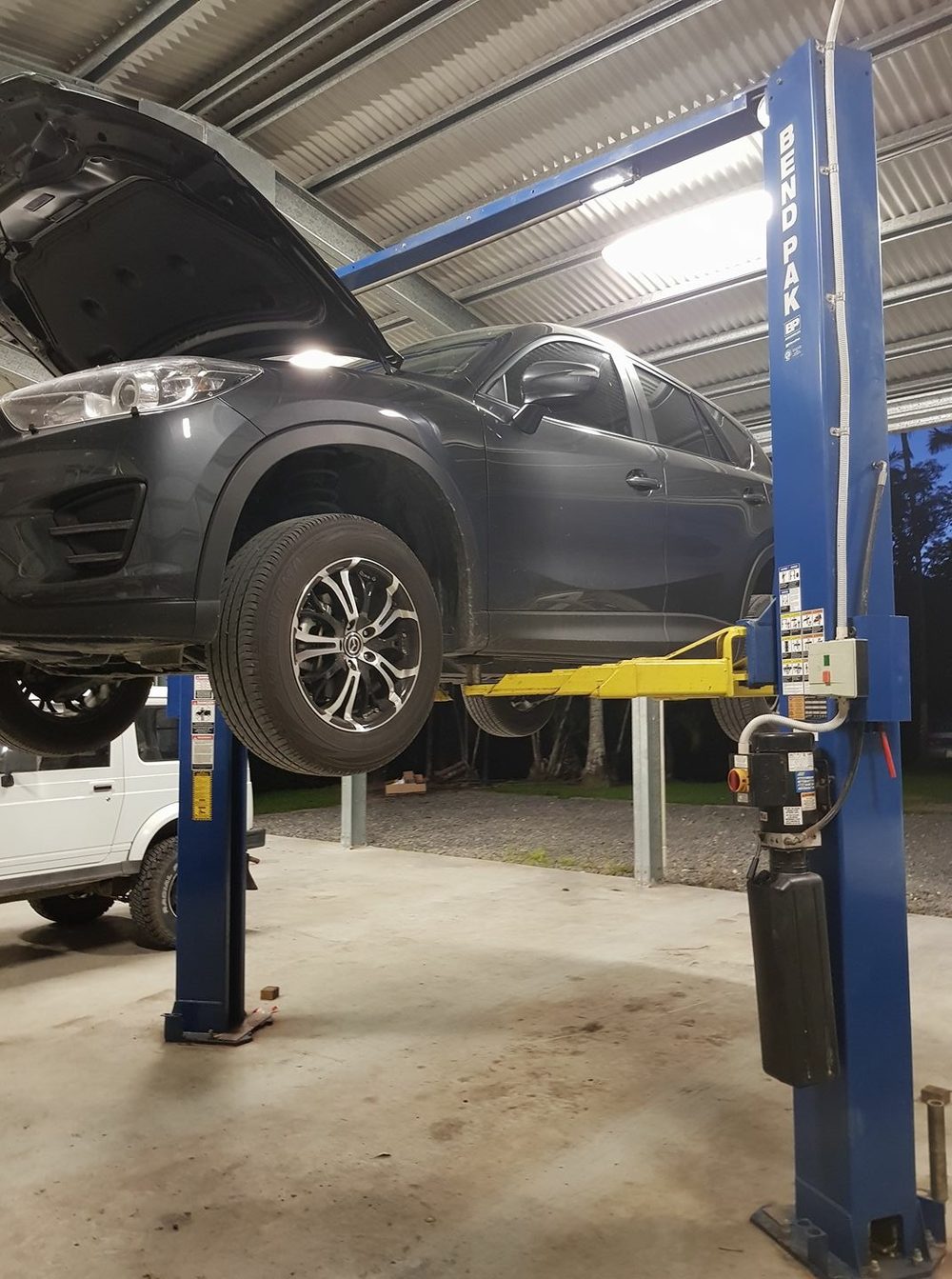 A car raised on the blue two post hoist at Mossman Mechanical in the evening, workshop lights on and palm trees outside